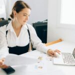 Woman at desk reviewing business reports on laptop, holding papers in a modern office setting.